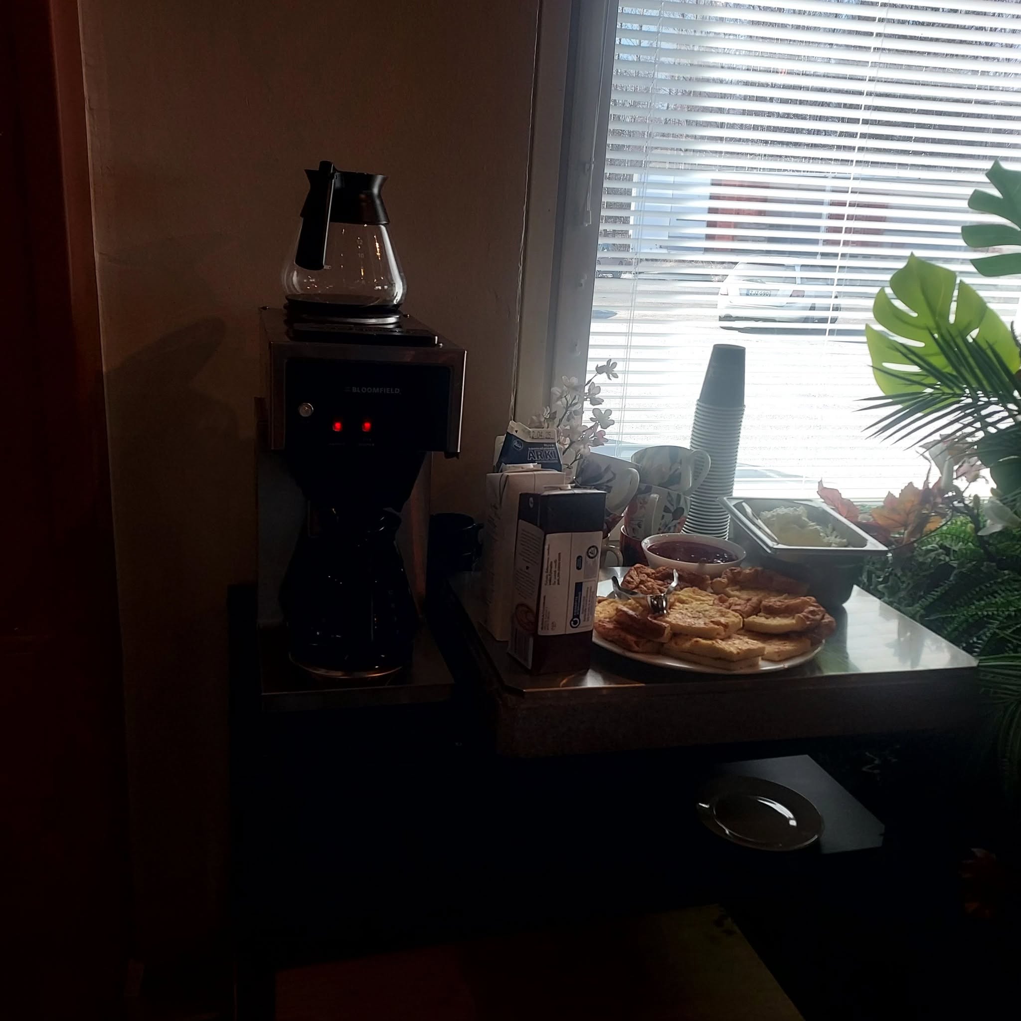 Kahvipöytä ikkunan äärellä: kahvinkeitin, pinot mukeja, leivonnaisia ja hilloa.